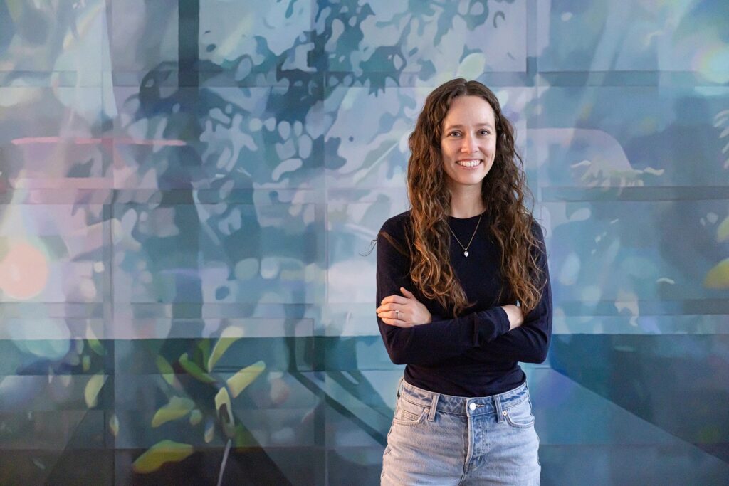 A person stands with folded arms before a layered blue-toned artwork, where soft shapes and shadows drift across the wall like reflected light.