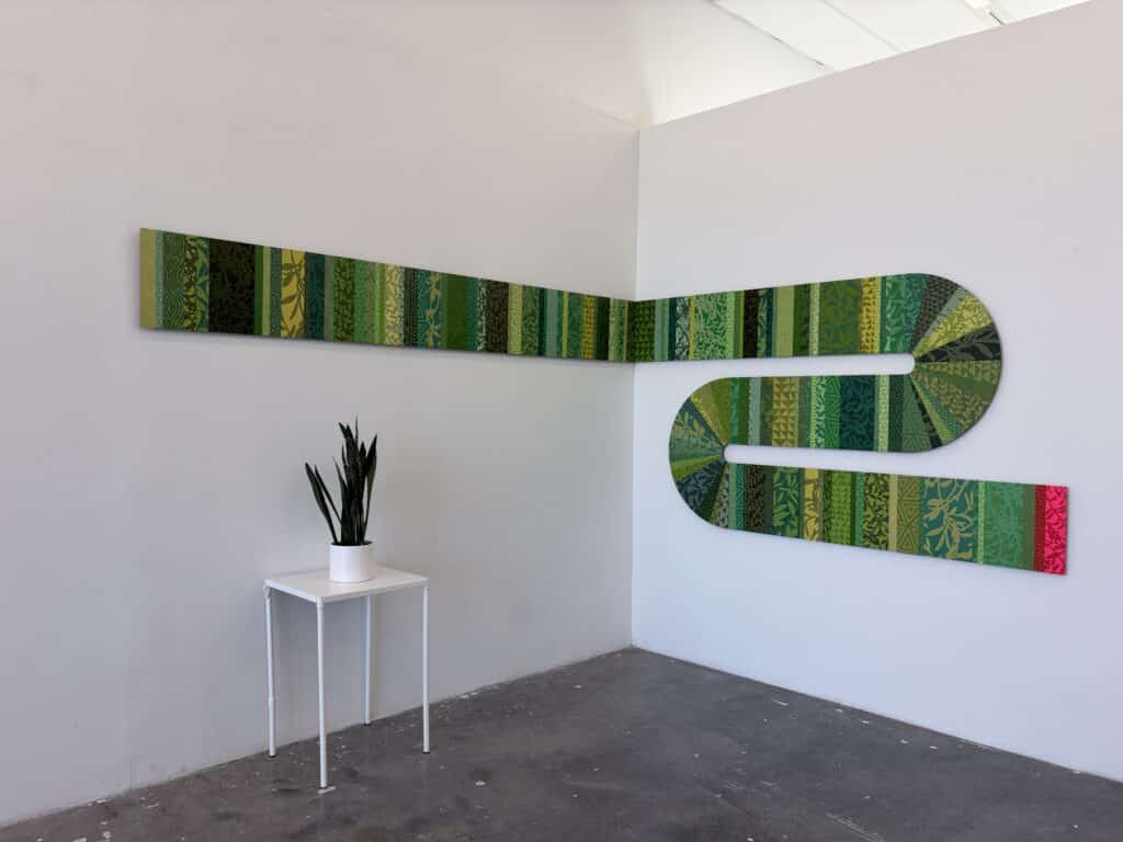 2026-MG-T-Anthropocene-in-situ-mid2-original_photo-1024x768 A long green artwork wraps around the corner of a white gallery wall, weaving together layered botanical patterns and geometric motifs. A small potted plant on a white table echoes the work's organic palette.