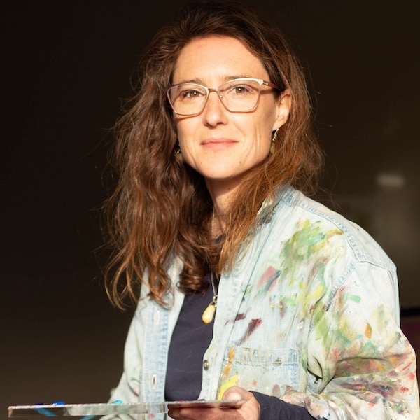 Sara Wiens. (Photo by Cindy Ursulak / courtesy Sara Wiens) An artist stands in warm studio light wearing a paint-splattered jacket and holding a palette, with a calm expression that reflects a creative practice in progress.